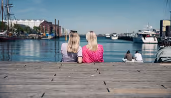 Två kvinnor som sitter på en träbrygga vänd mot en vattenmassa med båtar i bakgrunden. Dagen är klar och solig, med en blå himmel ovanför. Människor sitter i närheten och industribyggnader syns över vattnet.
