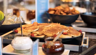 En frukostbuffé med ett fat med pannkakor toppat med jordgubbar och hallon. I närheten finns burkar med sirap och grädde, med fler frukostartiklar i bakgrunden, allt på en mörk disk.