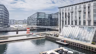 Modern scen vid vattnet med byggnader i glas som reflekterar solljus, en liten röd och vit fyr och båtar förlagda vid kanalen. En gångbro korsar vattnet under en delvis molnig himmel.