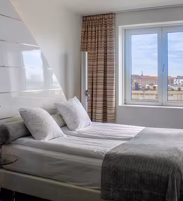 Modernt hotellrum med en prydligt bäddad dubbelsäng, vita sängkläder och grå filt. Det finns randiga gardiner, ett stort fönster med utsikt över staden och en röd stol bredvid en golvlampa. Atmosfären är ren och ljus.