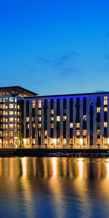 Modern byggnad med stora fönster upplysta på natten och reflekterar över en lugn vattenmassa. Himlen är mörkblå, vilket ger kontrast till det varma skenet från byggnaden.