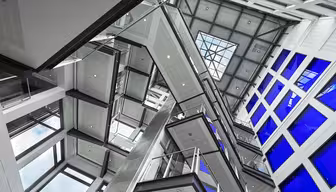 Interiörvy av en modern byggnad med flera trappor och balkonger i ett atrium. Designen har glas- och metallelement, med ett takfönster upptill. Stora blå fönster ger kontrast och skapar en öppen och futuristisk atmosfär.
