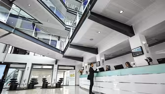En modern kontorslobby med en vit reception. Två sittande receptionister hjälper en stående person. Lokalen har högt i tak, ett glasatrium, flera våningar med räcken och modern inredning, inklusive abstrakt konst och eleganta möbler.