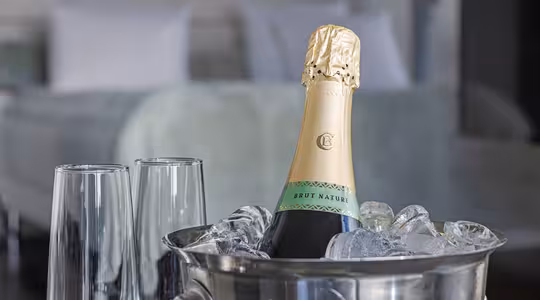 Champagneflaska i en ishink med två tomma flöjter bredvid, uppställd på ett bord. En suddig säng syns i bakgrunden, vilket tyder på en hotellrumsinställning.