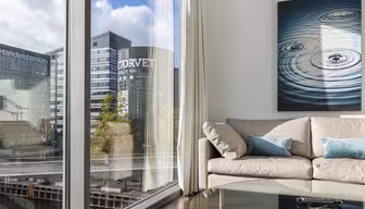 Ett modernt vardagsrum med en beige soffa och krickfärgade kuddar. På väggen hänger en målning av vattenkrusningar. Stora fönster ger utsikt över stadsbyggnader och en kanal, med "Handelsbanken" och "NORVET DESIGN MALL" synliga på intilliggande strukturer.