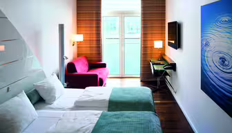 Ett modernt hotellrum med två enkelsängar, en röd fåtölj och en platt-TV på väggen. Det finns ett skrivbord med en stol bredvid TV:n. Blåvita sängkläder, randiga gardiner och en stor abstrakt målning syns.
