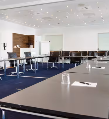 Konferensrum med långa svarta bord arrangerade i U-form, omgivet av svarta stolar. Anteckningsblock och pennor läggs på borden. Whiteboards, ett blädderblock och ett område för förfriskningar syns i bakgrunden. Rummet har stark belysning.
