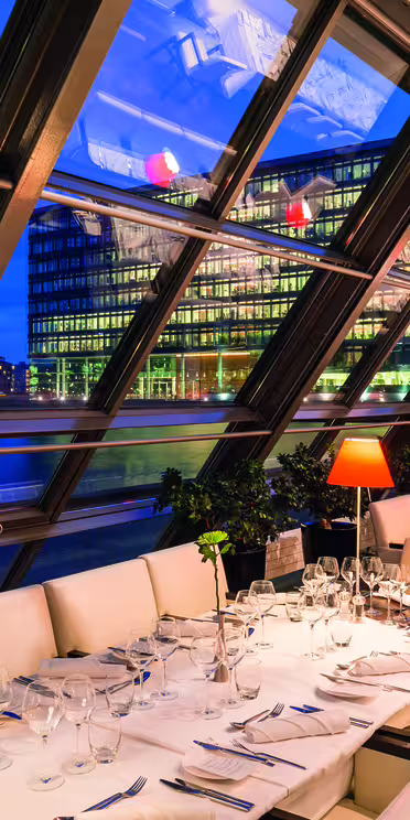 En modern restaurang med en glasvägg med utsikt över en stadsbild vid vattnet. Interiören har vita vadderade stolar, snyggt dukade bord med vinglas och röda bordslampor. Skylinen reflekteras på glaset, vilket ger en dynamisk stadsvy.