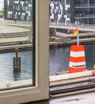 Utsikt från ett fönster som visar en kanal med en stor röd- och vitrandig boj med en flagga på toppen. Människor går och cyklar på den moderna strandpromenaden, och byggnader kantar den motsatta sidan av kanalen.